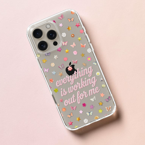 A clear iPhone case with a gray back, decorated with pastel pink and multicolor stars, butterflies, and the phrase 'everything is working out for me' in pink script, shown on a light pink background. (Feat)