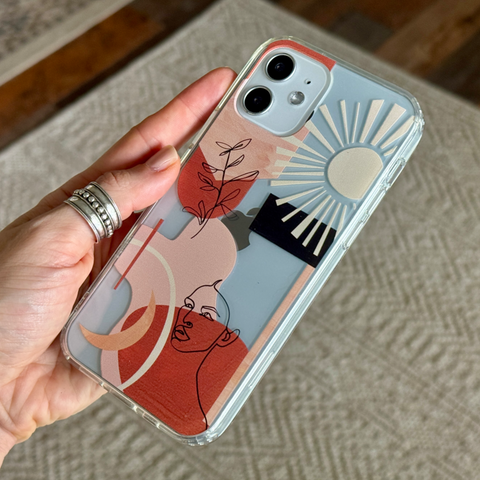 A hand holding a clear phone case decorated with an abstract art print in terracotta, dusty pink, cream, and black, set against a neutral textured rug. (Feat)