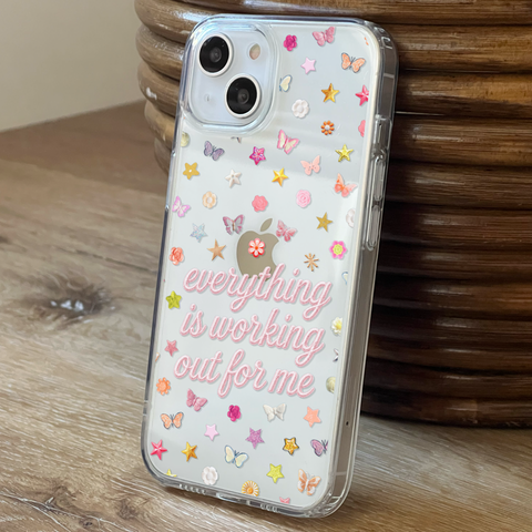 A clear phone case with a printed message and colorful printed embellishments, shown on an iPhone resting on a wooden surface next to a wooden basket. (Feat)