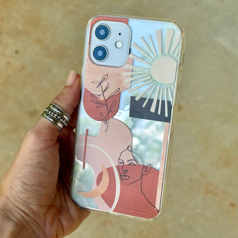 A hand holding a clear phone case with a colorful abstract art print featuring a sun, moon, and stylized face. The hand is adorned with silver rings, and the background is a soft-focus beige surface. (Feat)