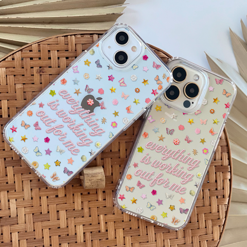 Two clear phone cases with pastel butterfly and star printed embellishments and the phrase 'everything is working out for me' in pink script, displayed on a woven round tray with neutral props. (Feat)