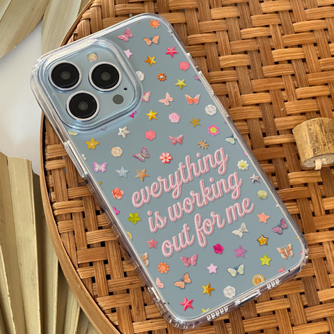 A clear phone case with a light blue back panel, decorated with colorful butterflies, stars, and the phrase 'everything is working out for me' in pink script, shown resting on a woven brown basket. (Feat)