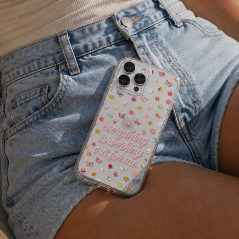 A clear phone case with colorful star and butterfly printed embellishments and the phrase 'everything is working out for me' in pink script, shown on a phone resting on a person's hip wearing denim shorts. (Feat)
