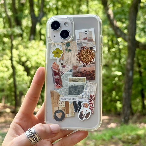 A hand holds a clear phone case decorated with vintage-style collage elements, including newspaper clippings, small flowers, and a circular mandala, against a blurred green forest background. (Feat)