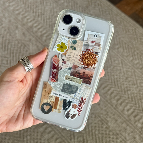 A hand wearing silver rings holds a clear phone case decorated with a vintage-style collage of paper clips, stamps, a small yellow flower, and a woven basket motif, against a textured beige background. (Feat)