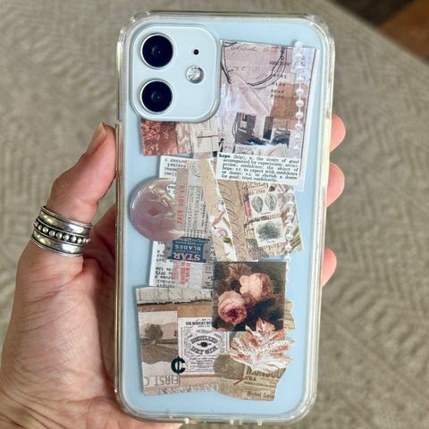 A hand holding a clear phone case, decorated with vintage newspaper clippings, small floral prints, and a pink stone accent. (Feat)