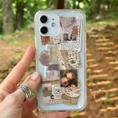A hand holds a clear phone case decorated with vintage-style collage elements, including newspaper clippings, floral prints, and a pink stone, against a blurred outdoor forest background. (Feat)