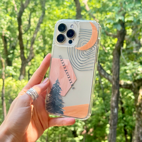 A hand holding a clear iPhone case decorated with a minimalist landscape design featuring a crescent moon, pine tree, and geometric shapes in peach, navy, and gray, set against a blurred green forest background. (Feat)