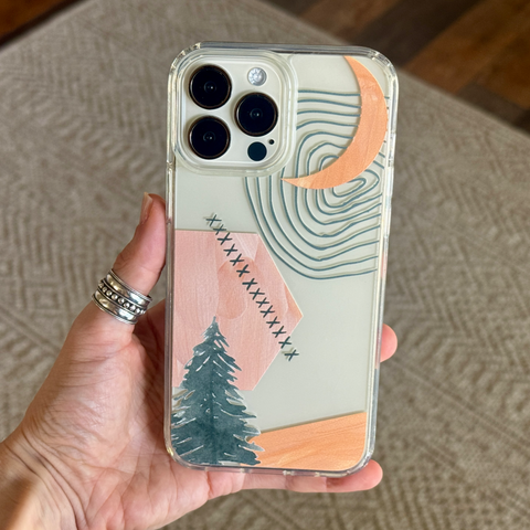 A hand holds a clear phone case featuring a minimalist landscape design with a pine tree, pink and peach geometric shapes, and teal contour lines on a light background. (Feat)