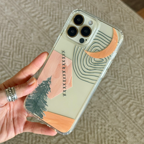 A hand holding a clear phone case decorated with a minimalist landscape design featuring a pink house, pine tree, moon, and topographic lines on a light beige background. (Feat)