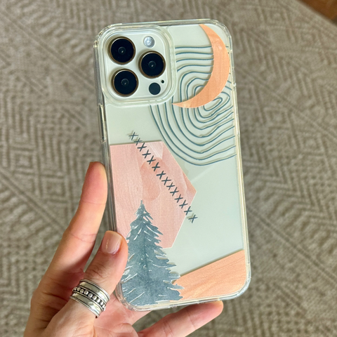 A hand holding a clear phone case decorated with a minimalist landscape design featuring a pine tree, pink mountain, blue waves, and a peach crescent moon, set against a textured gray background. (Feat)