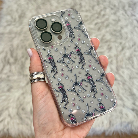 A hand holds a clear phone case decorated with black skeleton dancers and small pink and silver rhinestone accents, shown against a soft white faux fur background. (Feat)