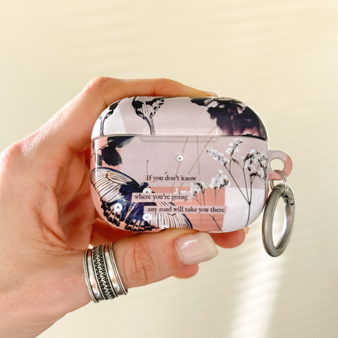 A hand wearing silver rings holds an AirPods case decorated with a vintage-style floral and butterfly print in pink, black, and white, complete with a silver keychain ring. (Feat)
