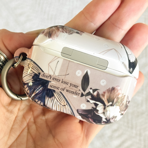 A hand holding a white AirPods case decorated with a vintage-style floral and butterfly print in black, beige, and navy, featuring the quote 'don't ever lose your sense of wonder' on the side.  (Feat)