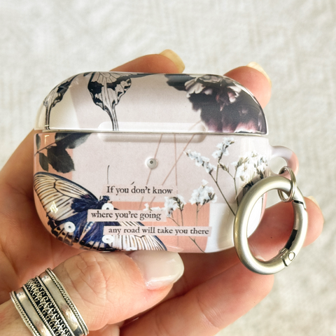 A hand with silver rings holds a decorative AirPods case featuring a floral and butterfly pattern in pink, black, and white, with a silver keychain ring attached.  (Feat)