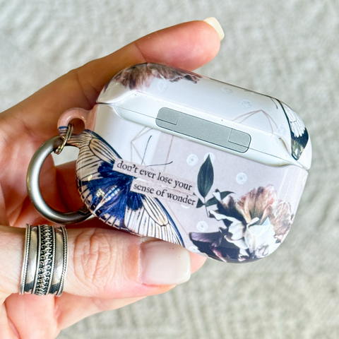 A hand holding a white AirPods case decorated with a blue and brown butterfly print, floral motifs, and the quote 'don't ever lose your sense of wonder' in black script. The case has a silver metal keychain ring attached.  (Feat)