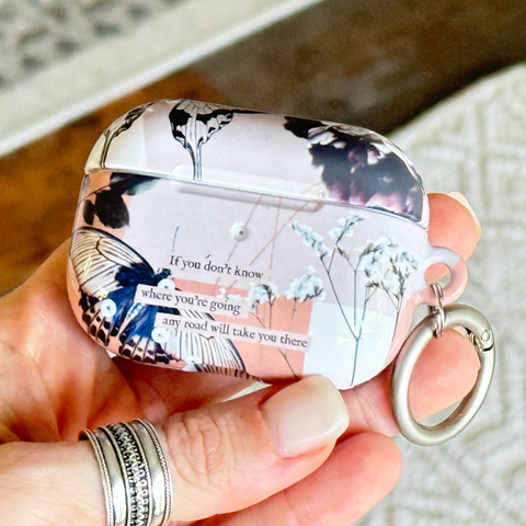 A hand holds a protective AirPods case with a floral and butterfly print, featuring a silver metal key ring attached.  (Feat)