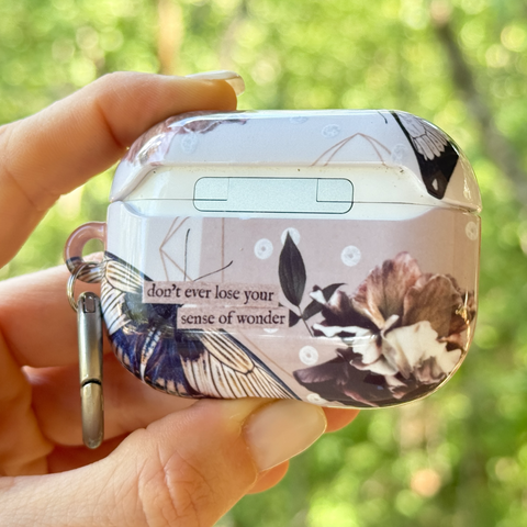 A hand holding a protective case for an AirPods charging case, decorated with a vintage-style floral and butterfly print in muted brown, beige, and navy tones, with a silver keychain attached. The background is a soft-focus green outdoor setting.  (Feat)