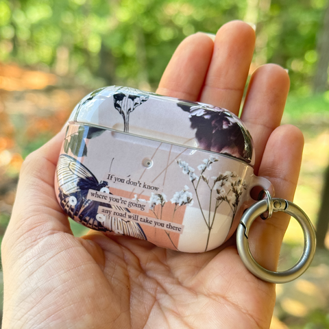 A hand holds a decorative AirPods case with a butterfly and floral design, a printed quote, and a silver key ring attachment, set against a blurred green forest background.  (Feat)